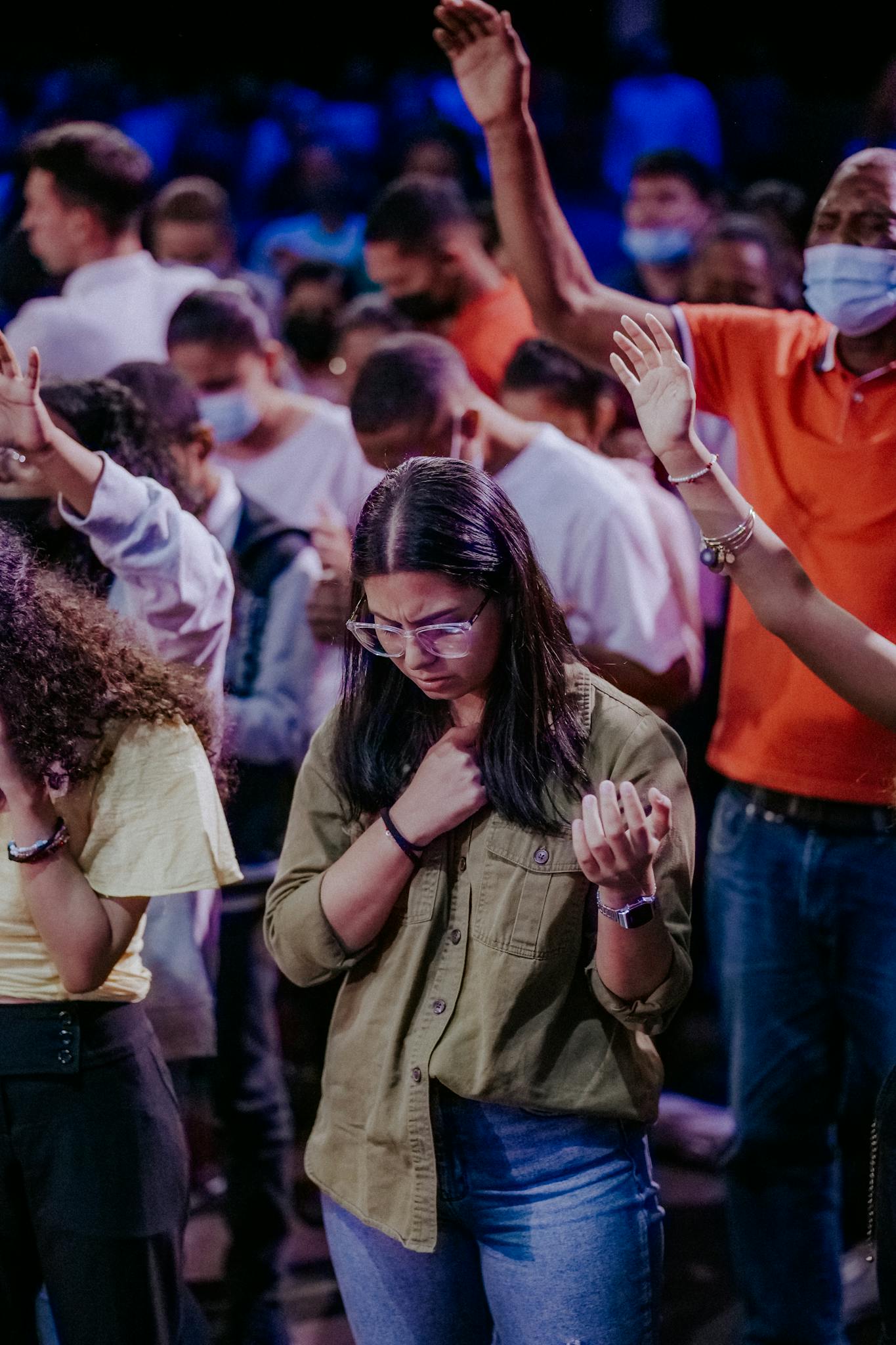 A diverse crowd participates in an indoor reflective gathering with hands raised.